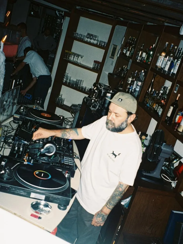 Şapkalı, sakallı ve kolları dövmeli bir DJ, arkasında çeşitli içki şişeleri ve kadehlerin sıralandığı ahşap rafların bulunduğu loş ışıklı bir tavernada, pikaplar ve mikser başında müzik seçip çalarken canlı bir atmosfer yaratıyor.