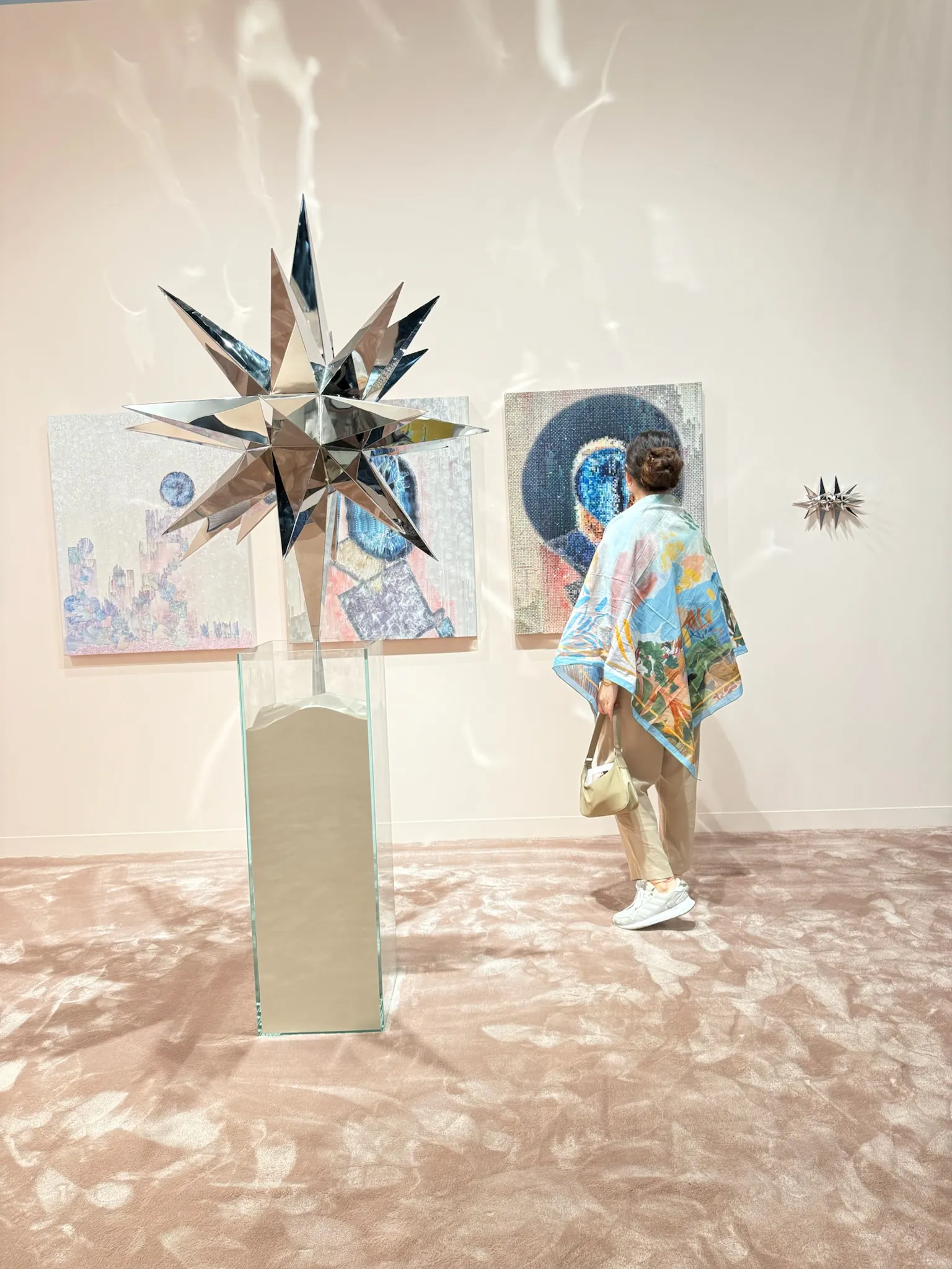 Abu Dhabi Art’ta ayna yüzeyli yıldız formunda bir heykel ve arka planda çağdaş resimlerin yer aldığı sergi alanını inceleyen ziyaretçi