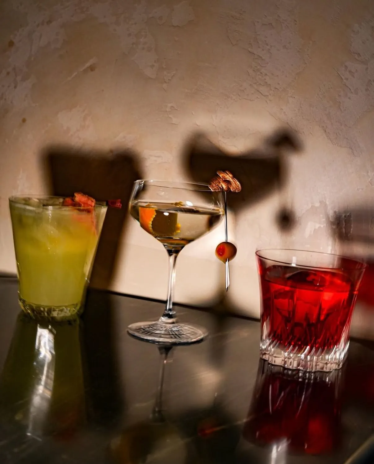 Ambiyanslı bir bar ortamında, koyu renkli yansıtıcı bir yüzey üzerinde dizili, her biri farklı renk ve garnitürlerle özenle hazırlanmış üç kokteyl; arka plandaki dokulu duvarda oluşan belirgin gölgeler ve sıcak ışıklandırma ile şık bir atmosfer sunuyor.