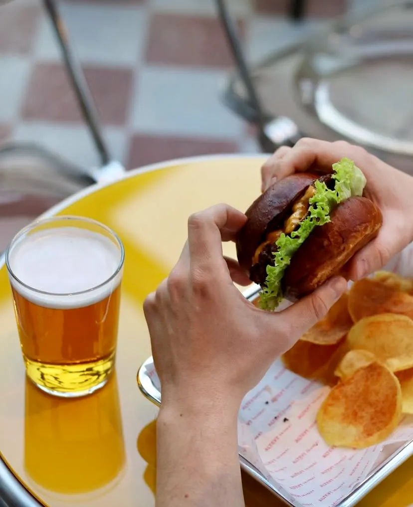 Alt-Kat Drinkery'nin canlı ortamında, parlak sarı bir masanın üzerinde duran köpüklü bir bira bardağının yanında, bir elin taze marul, erimiş peynir ve bol soslu bir burgeri tuttuğu, çıtır patates cipsleriyle zenginleşen lezzetli bir an.