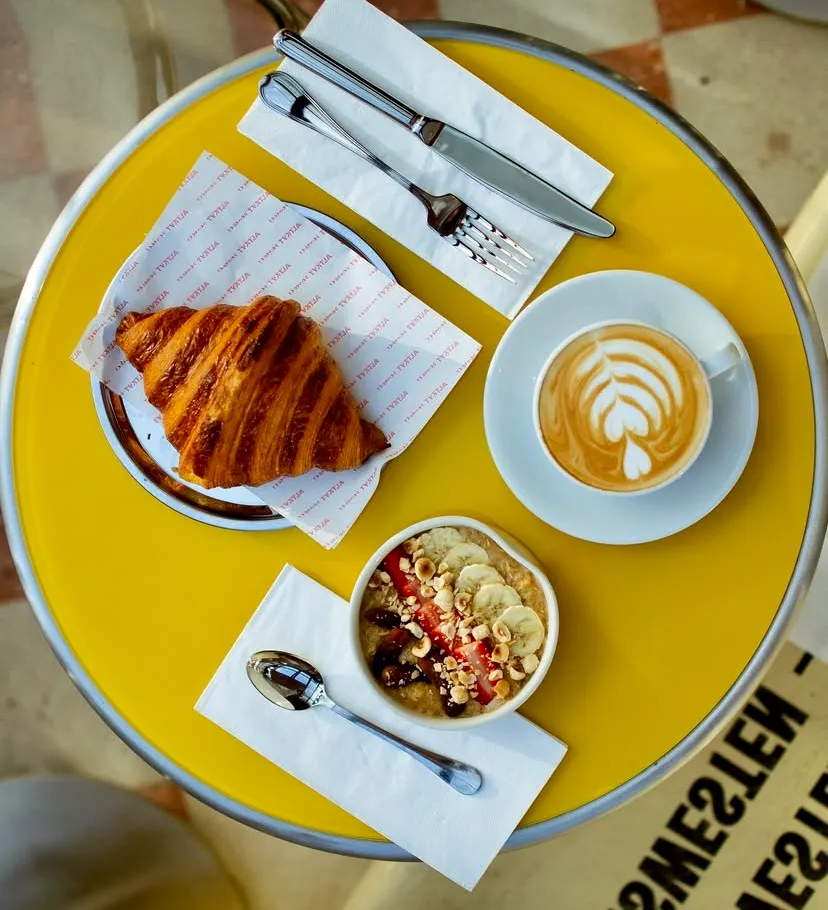 Parlak sarı yuvarlak bir masanın üzerinde, Alt Kat Drinkery'nin özenle hazırladığı, çıtır altın rengi bir kruvasan, kalpli latte sanatına sahip aromatik bir kahve ve muz, çilek, kıyılmış fındıkla süslenmiş besleyici bir yulaf ezmesi kasesi sergileniyor.