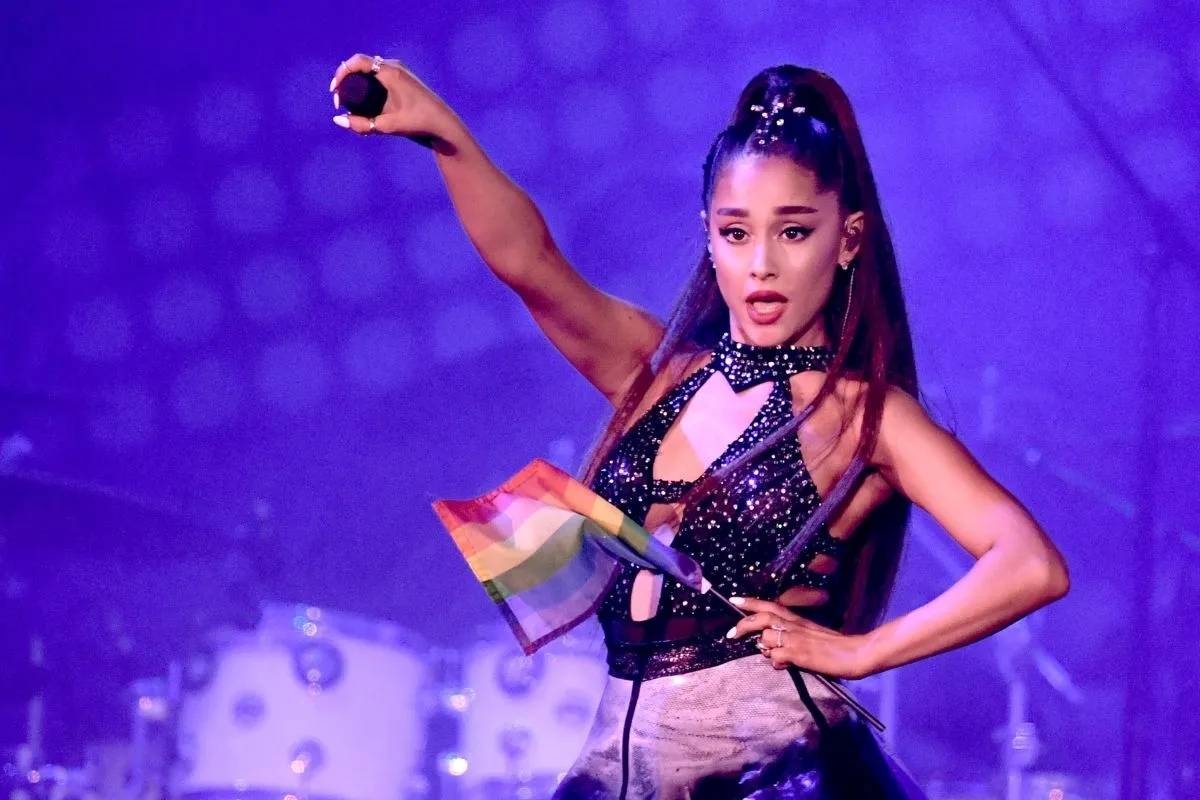 Ariana Grande, mor ve mavi ışıklarla aydınlatılmış sahnede, yüksek at kuyruğu ve parlak detaylı kostümüyle şarkı söylerken, bir elinde mikrofonu havaya kaldırmış, diğer elinde küçük bir gökkuşağı bayrağı tutarak büyük müzik festivallerinin ruhunu yansıtan enerjik bir performans sergiliyor.