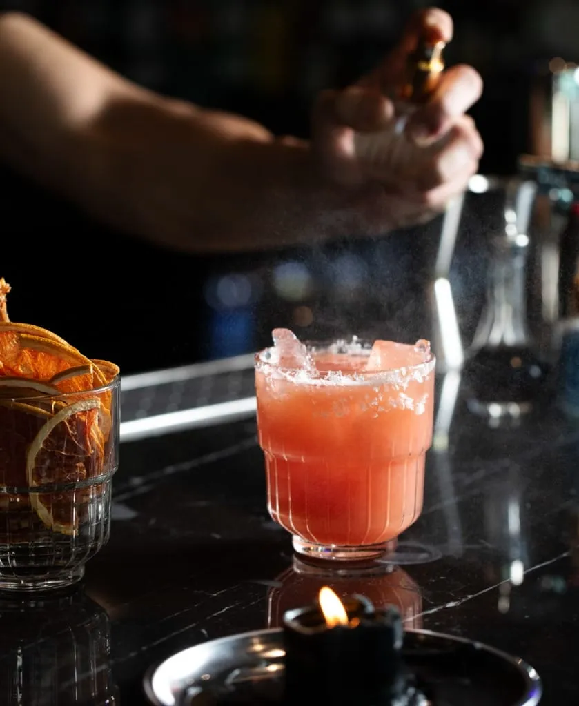 Barmen, karanlık bir bar ortamında, parlak siyah mermer tezgah üzerindeki tuz kenarlı, buzlu ve pembe renkli yivli bir kokteylin üzerine küçük bir şişeden aromatik sprey sıkarken, hemen yanında kuru portakal dilimleriyle dolu bir bardak ve yanan bir mum bu özel anın atmosferini tamamlıyor.