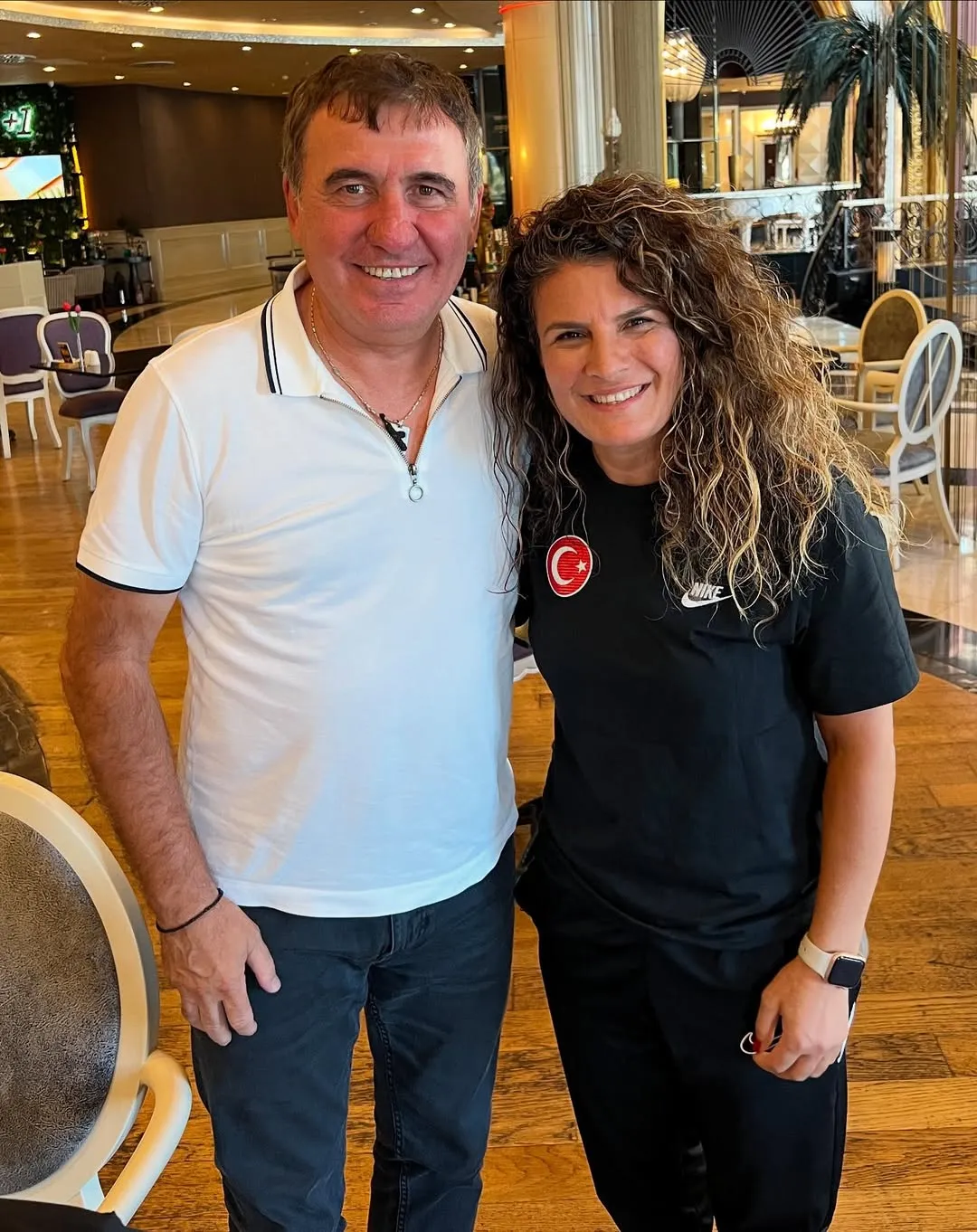 Kadın futbolcu Bilgin Defterli ile beyaz polo tişörtlü bir erkek, lüks bir iç mekanda neşeyle gülümseyerek kameraya bakıyor; Bilgin Defterli'nin siyah tişörtünde Türk bayrağı arması ve Nike logosu öne çıkıyor.