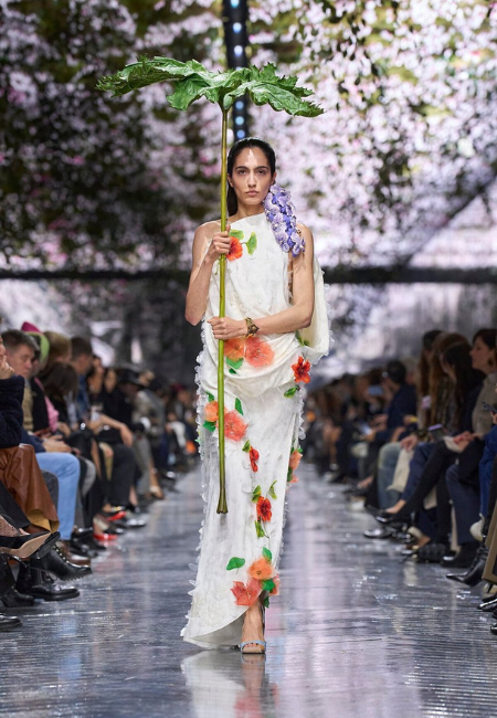 Paris 2026 Couture Moda Haftası’nda Dior haute couture defilesinden çiçek aplikeli beyaz elbise ve botanik temalı dramatik podyum görünümü.