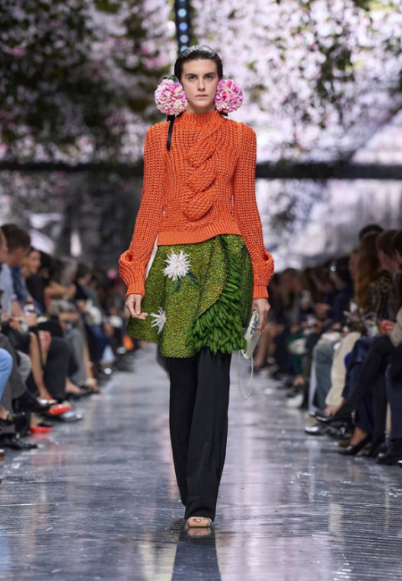 Dior 2026 haute couture defilesinde turuncu örgü kazak ve yaprak dokulu yeşil etekle sunulan doğadan ilham alan podyum görünümü.