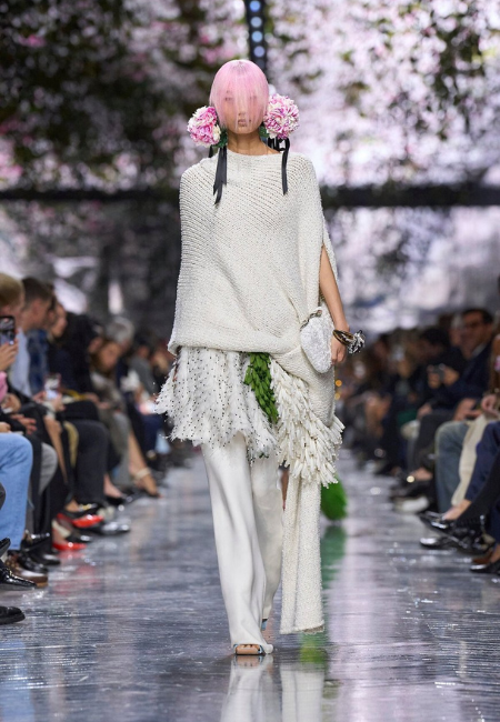Dior 2026 haute couture defilesinde beyaz katmanlı elbise, el işçiliği dokular ve floral saç aksesuarlarıyla sunulan podyum görünümü.
