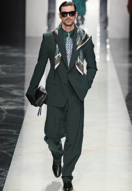 Dolce&Gabbana 2026 erkek moda haftası yeşil takım elbise, desenli kravat, omuzda şal detaylı aksesuar ve clutch çanta ile şık görünüm.