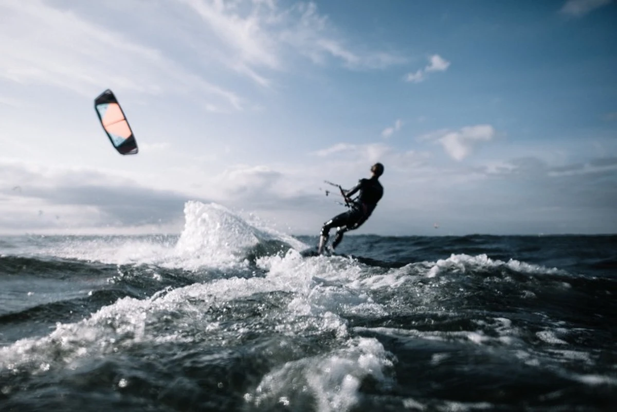 Mavi ve beyaz bulutlu gökyüzünün altında, siyah bir wetsuit giyen bir kitesurfçü, dalgalı denizde hızla ilerlerken renkli uçurtmasıyla suda büyük bir köpük patlaması yaratıyor, en iyi sörf noktalarından birindeki dinamik ve heyecan verici bir anı yansıtıyor.