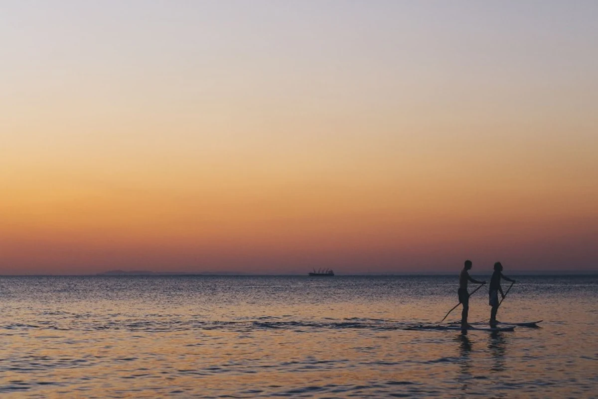 Gün batımının turuncu ve pembe tonlarıyla gökyüzünün yansıdığı sakin denizde, iki kişinin stand-up paddleboard ile kürek çektiği, Silivri gibi en iyi SUP noktalarının keyfini çıkardığı huzurlu bir an tasvir ediliyor.