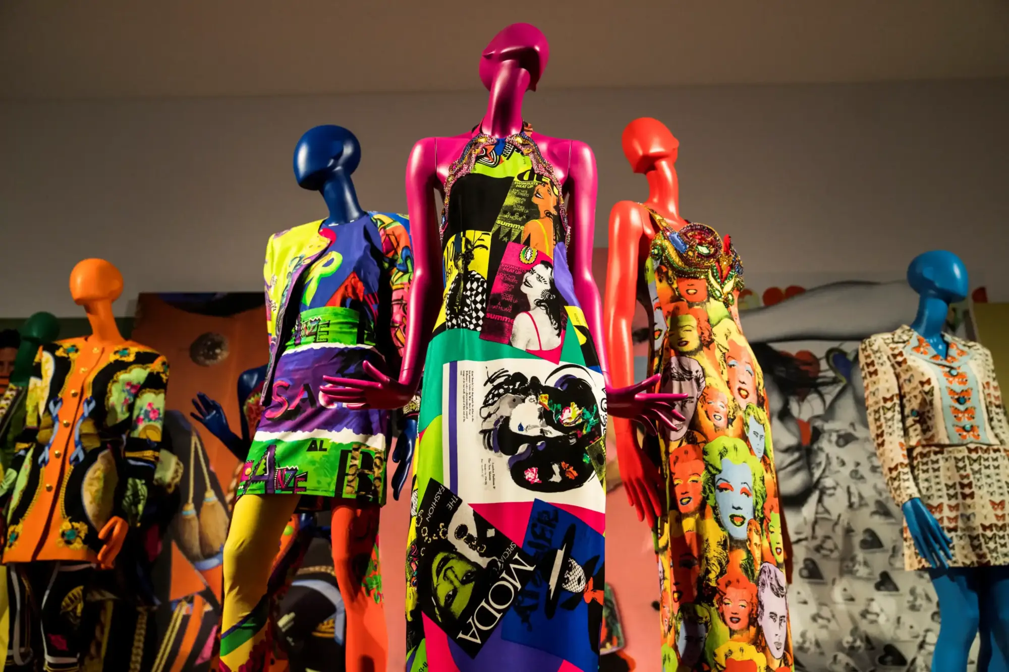 2026 yılındaki dünya çapındaki moda sergileri rehberine eşlik eden bu görselde, pop art ve 90'lar esintileri taşıyan, Marilyn Monroe yüzleri ve dergi kupürleri gibi canlı desenlerle süslü, pembe, mavi, turuncu ve kırmızı renkli mankenler, modanın enerjik dönüşümünü ve sanatsal ifadesini yansıtıyor.