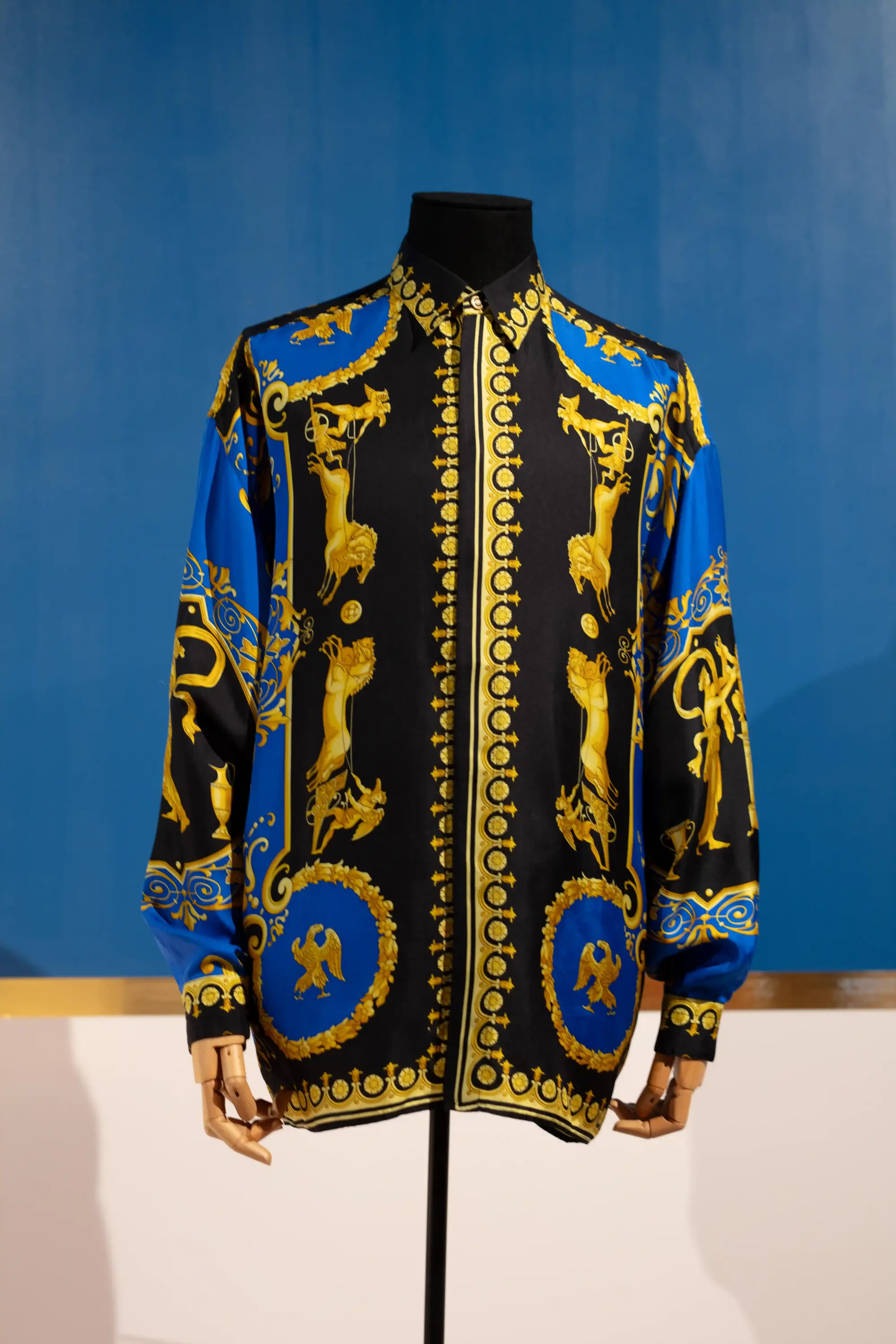 Manken üzerinde sergilenen siyah zemin üzerine altın ve mavi barok desenlerle bezeli, Versace'ye özgü gösterişli uzun kollu bir gömlek, 2026 yılındaki uluslararası moda sergileri kapsamında modanın zanaat ve tasarım yönünü vurguluyor.