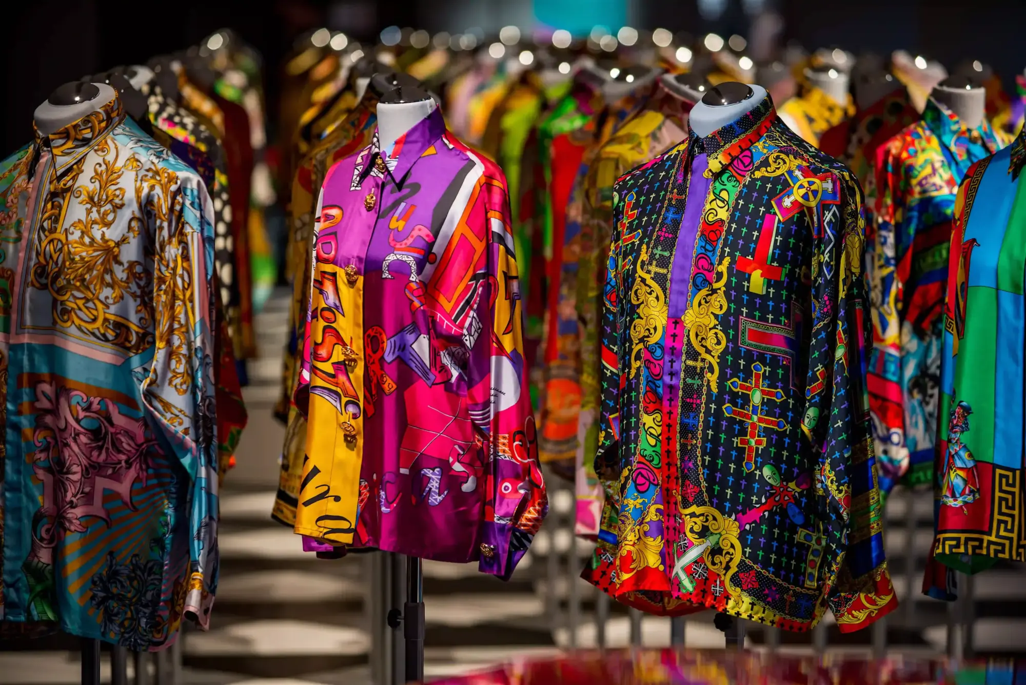 Koyu fon üzerinde sıralanmış, canlı mor, fuşya, sarı, siyah, altın, kırmızı ve turkuaz renklerde barok ve pop art desenlerle süslenmiş, ikonik Gianni Versace imzasını taşıyan ipek gömlekler, dünya moda sergileri kapsamında sergileniyor.