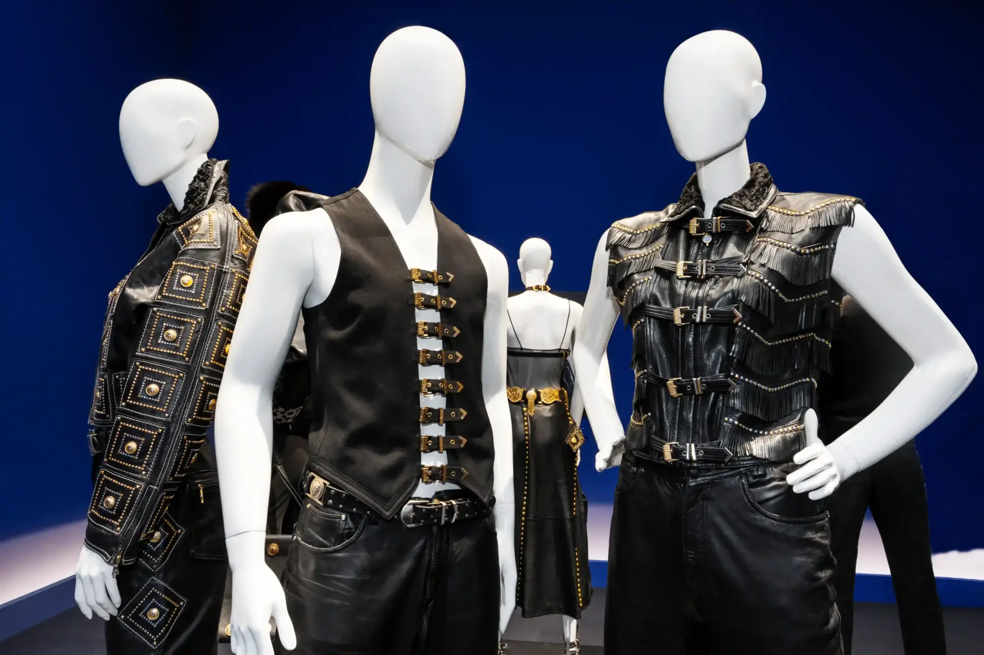 Bir moda sergisinde, koyu mavi arka plan önünde, altın rengi zımbalar, tokalar ve püsküllerle süslenmiş siyah deri ceket, yelek ve pantolon giymiş cansız mankenler, Gianni Versace'nin ikonik ve gösterişli tasarımlarını sergiliyor.