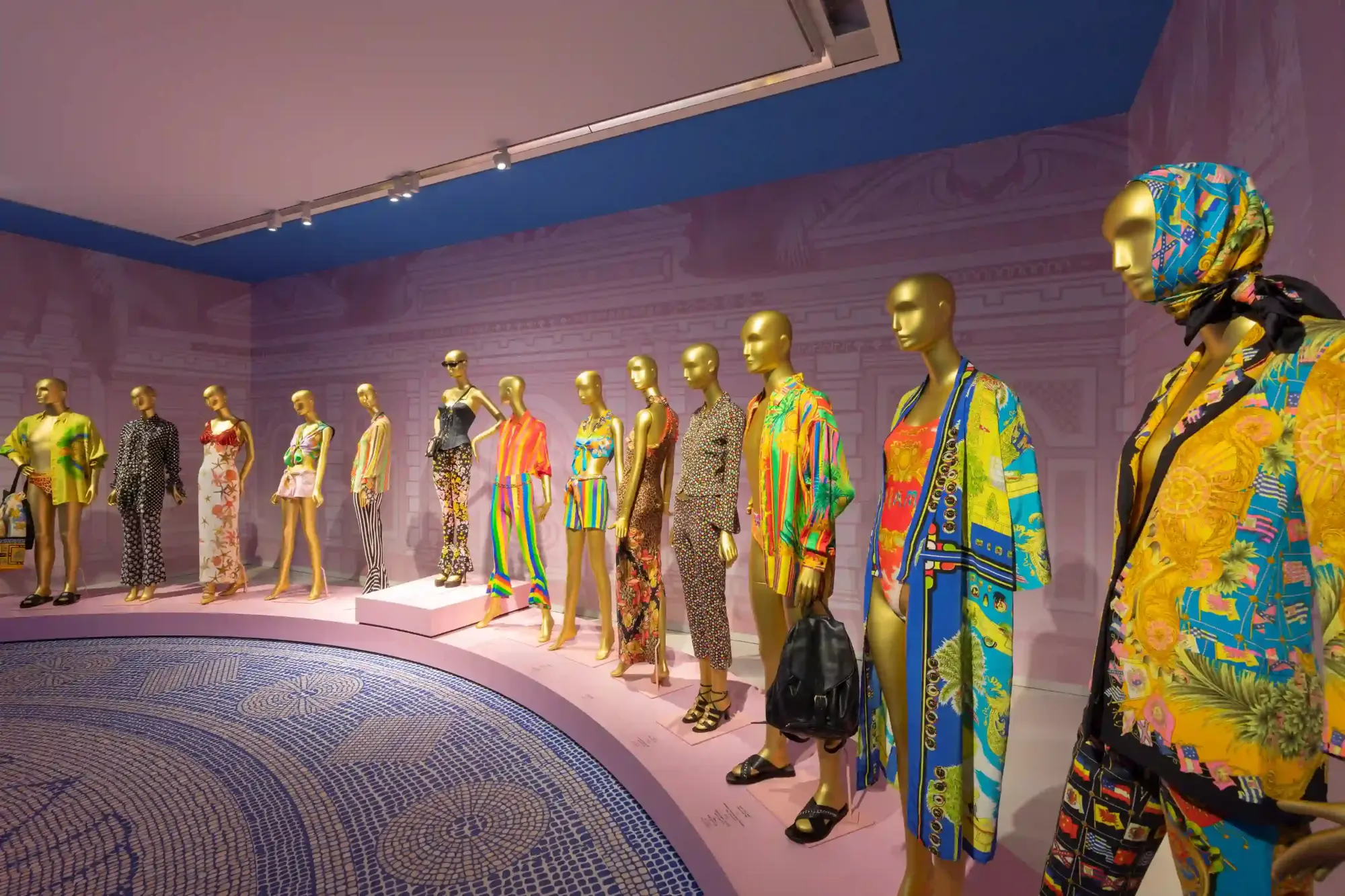 Bir moda sergisi alanında, altın renkli mankenler üzerinde tropikal motifler, barok süslemeleri andıran altın detaylar ve canlı şeritli desenlerle zenginleştirilmiş iddialı elbiseler, plaj giysileri ve ipek eşarplar sergileniyor; pembe tonlardaki dekorasyon ve alttaki karmaşık mavi mozaik zemin bu gösterişli koleksiyonu vurguluyor.