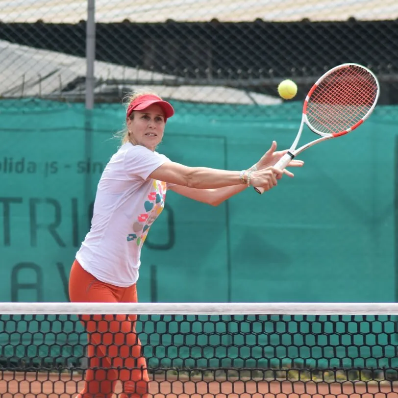 İpek Şenoğlu, kırmızı vizörü, renkli desenli beyaz tişörtü ve turuncu taytıyla bir tenis kortunda, topa vurmak için raketini çift elle sıkıca tutarken, sarı bir tenis topu raketinin hemen üzerinde havada asılı kalmış, file ve yeşil kort zeminli bir arka plan önünde dinamik bir anı yakalıyor.