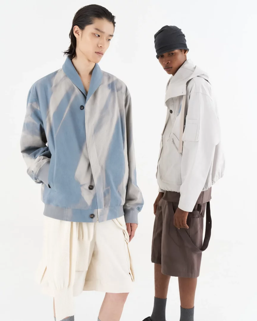 Pitti Uomo'da yeni nesil erkek modasının deneysel ve yenilikçi kodlarını yansıtan bir görsel: Jiyong Kim'in güneş ışığında doğal olarak soldurulmuş, açık mavi ve gri tonlarındaki bomber ceketiyle Asyalı bir model ve kapüşonlu bej bir ceketle kahverengi şort giyen siyahi bir model yan yana duruyor.