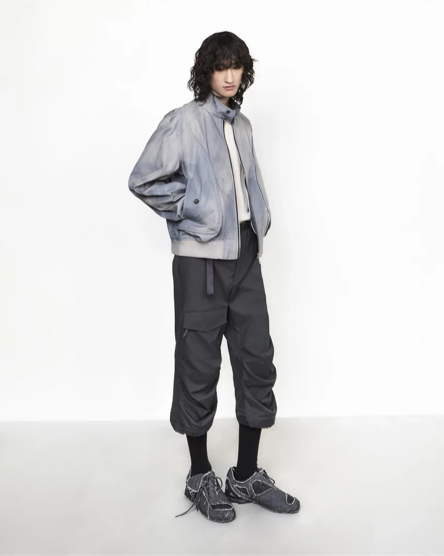 Jiyong Kim'in Pitti Uomo'da sergilediği koleksiyondan, güneş ışığında doğal olarak soldurulmuş açık mavi bomber ceket ve bol paçalı kapri pantolon giyen model, erkek modasında yenilikçi materyal kullanımını vurguluyor.