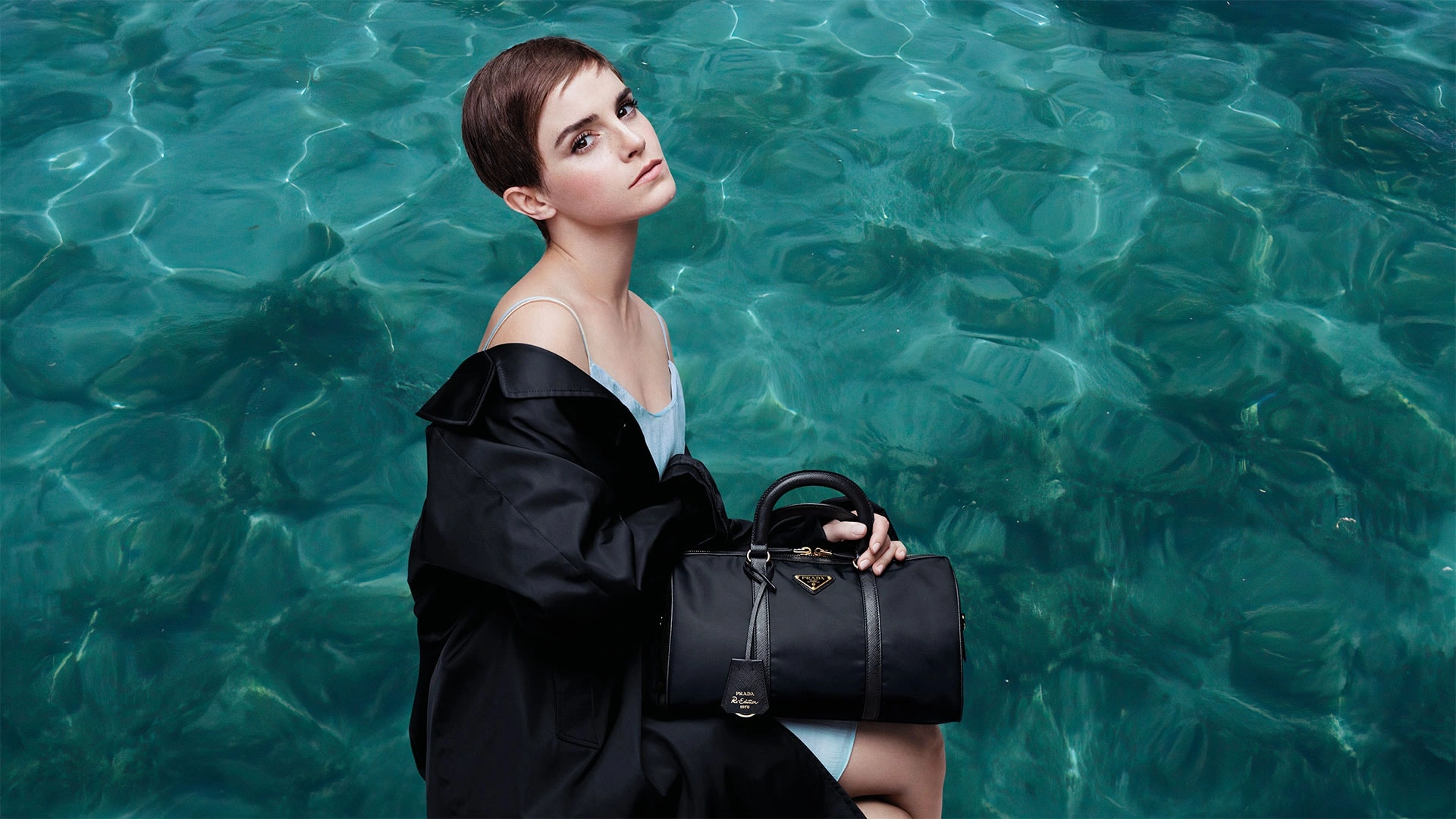 Turkuaz deniz fonunda siyah Prada çanta taşıyan model; modern lüks estetiği, zamansız stil ve karbon nötr moda yaklaşımını yansıtan kampanya görseli.