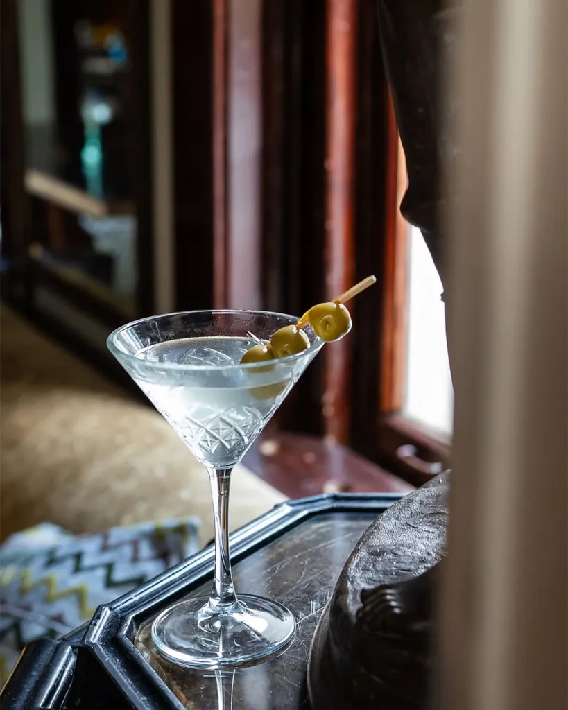 Zarif detaylara sahip kesme cam bir martini kadehinde, üç yeşil zeytinle süslenmiş berrak bir kokteyl, loş ışıklı şık bir iç mekanda, ahşap pencere çerçeveleri ve hafifçe perdelenmiş bir fon eşliğinde, İstanbul'un zengin kokteyl sahnesinden özel bir anı yansıtarak, rahatlatıcı ve stil sahibi bir kokteyl mekanı atmosferi sunuyor.