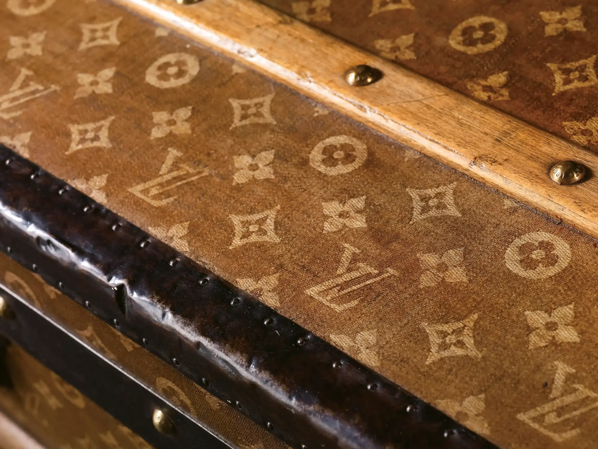 Kahverengi dokuma kanvas üzerine işlenmiş açık renk 'LV' monogram harfleri ve stilize çiçek motifleriyle bezeli, koyu renk deri ve pirinç detaylarla güçlendirilmiş eski bir Louis Vuitton sandığın üst yüzeyi, Georges Vuitton tarafından 1896'da tasarlanan bu ikonik desenin 130 yıllık köklü geçmişini ve zanaatkârlığını vurguluyor.