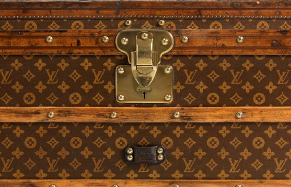 Eski bir Louis Vuitton sandığının yakın çekiminde, markanın 130 yıllık hikâyesini simgeleyen, iç içe geçmiş LV harfleri ve stilize çiçeklerden oluşan kahverengi ikonik Monogram deseni, pirinç çivilerle süslenmiş ahşap detaylar ve merkezi bir pirinç kilit mekanizması dikkat çekiyor.
