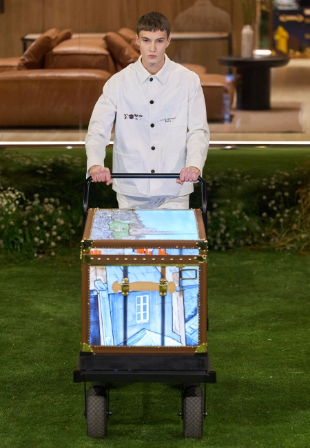 Louis Vuitton 2026 Sonbahar/Kış erkek koleksiyonunda beyaz ceketli model, Louis Vuitton sandık arabasıyla podyumda.