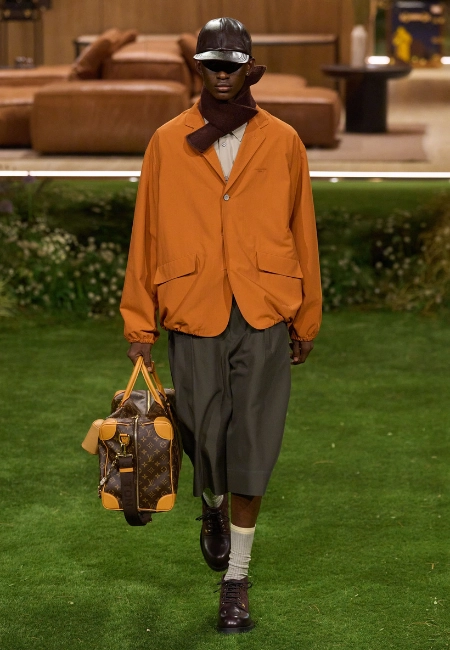Louis Vuitton 2026 Sonbahar/Kış erkek koleksiyonunda turuncu ceket, deri şapka ve monogram valiz çanta görünümü.