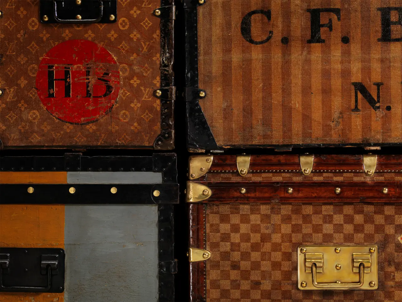 Eski Louis Vuitton sandıklarının yakın çekim görüntüsü, markanın ikonik Louis Vuitton Monogram deseni, çizgili ve Damier kanvas desenleri ile farklı renk panellerini bir araya getirerek, sandıkların yüzeyindeki detaylarla 130 yıllık zanaat ve tasarım hikayesini çarpıcı bir şekilde gözler önüne seriyor.