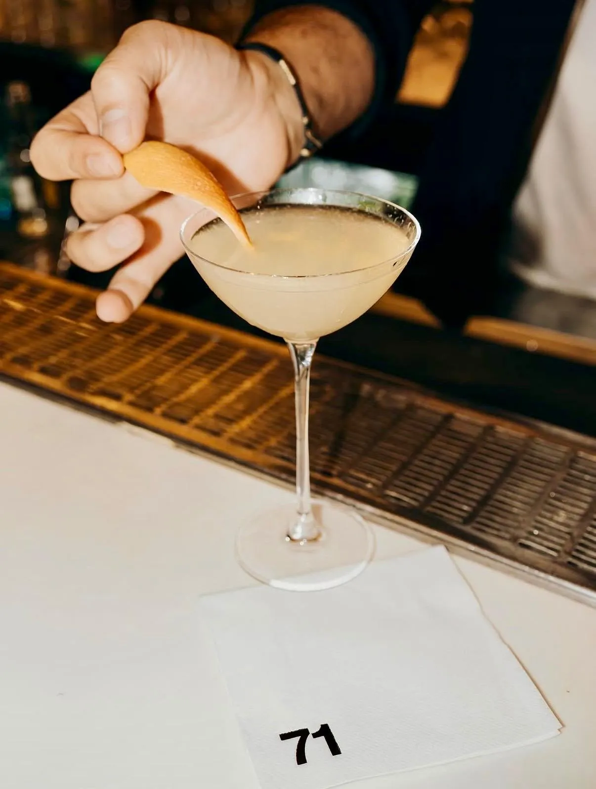 Barmen, Lucca Bebek atmosferine uygun, özel bir kokteylin sunumunu portakal kabuğu ile tamamlarken, parlak ışıkta parlayan içecek kadehi ve altında 71 yazılı peçete, mekanın şık detaylarını yansıtıyor.