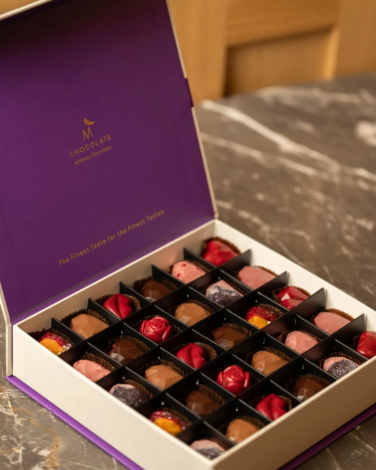 M Chocolate Artisan Chocolate imzalı, mor kutu içinde farklı dolgulara sahip el yapımı çikolata seçkisi.