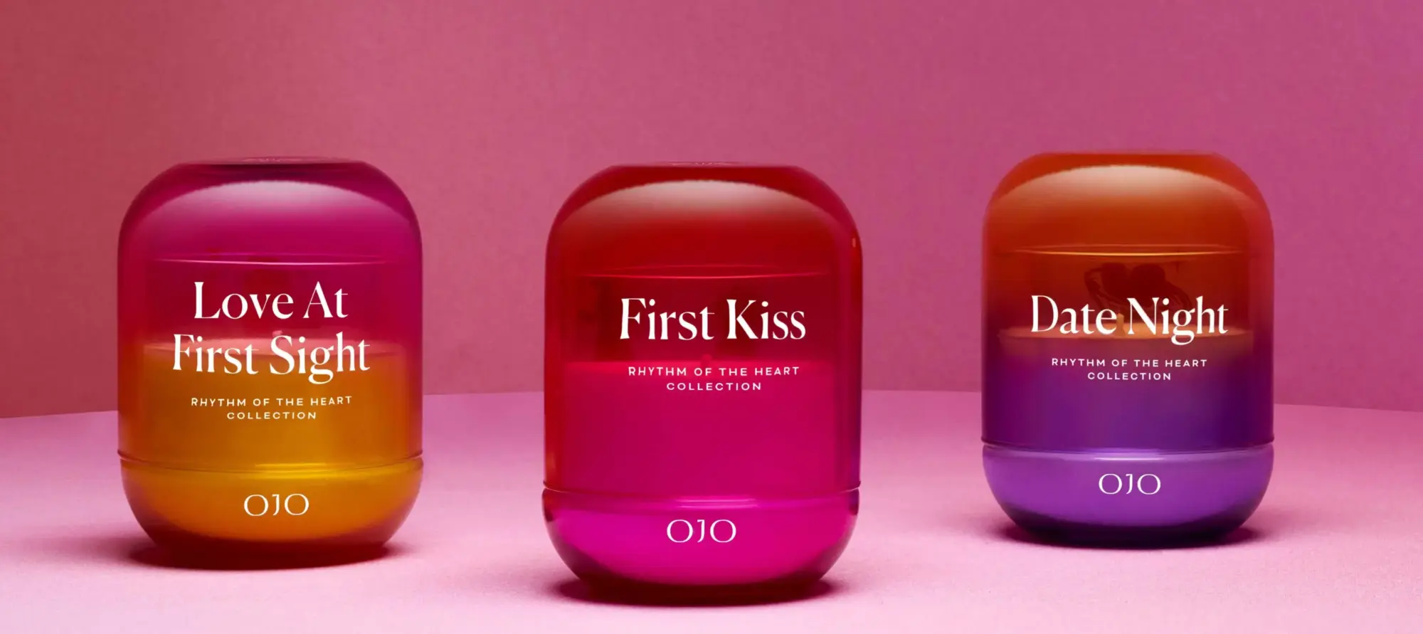OJO'nun Rhythm of the Heart Koleksiyonu'ndan üç kokulu mum, pembe zemin üzerinde yan yana dizilmiş. Çok renkli kapların üzerinde Love At First Sight, First Kiss ve Date Night isimleri yazıyor.