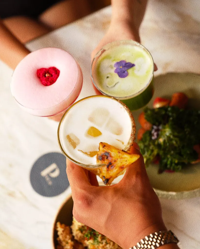 Keyifli bir ana tanıklık eden bu fotoğrafta, üç farklı kişinin Paolina Cocktail Bar'da çeşitli, özenle hazırlanmış kokteyllerle kadeh kaldırdığı an görülüyor: biri kalpli kırmızı bir garnitürle süslenmiş pembe köpüklü, diğeri mor çiçekli açık yeşil, diğeri ise ızgara ananas dilimiyle ferahlatıcı bir içecek.