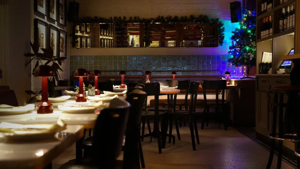Nişantaşı'nda yer alan, loş ışıklarla aydınlatılmış şık bir restoranın içi; kırmızı masa lambaları, yılbaşı süslemeleri, duvarlardaki çerçeveli resimler ve aynalarla davetkar bir atmosfer sunarken, arka plandaki mavi-mor ışıklı dekoratif karolar ve ışıklı bir yılbaşı ağacı mekana modern ve şenlikli bir hava katıyor.