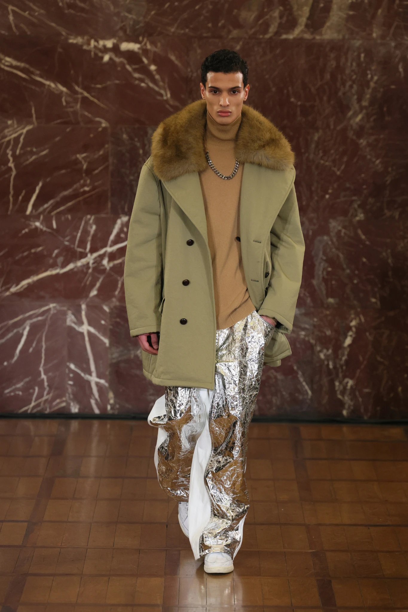 Hed Mayner'ın Pitti Uomo defilesinde bir model, kürklü bej palto, balıkçı yaka kazak ve parlak metalik gümüş pantolon giyerek mermer bir duvarın önündeki podyumda kararlılıkla yürüyor.