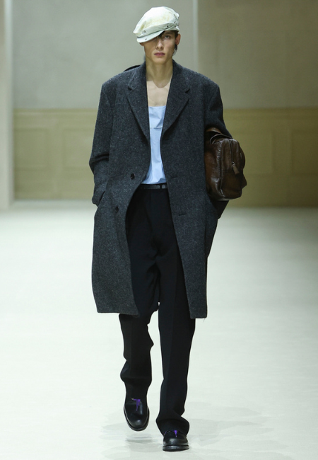 Prada 2026 menswear koleksiyonu gri yün palto, beyaz şapka ve siyah pantolon ile klasik şehir görünümü.
