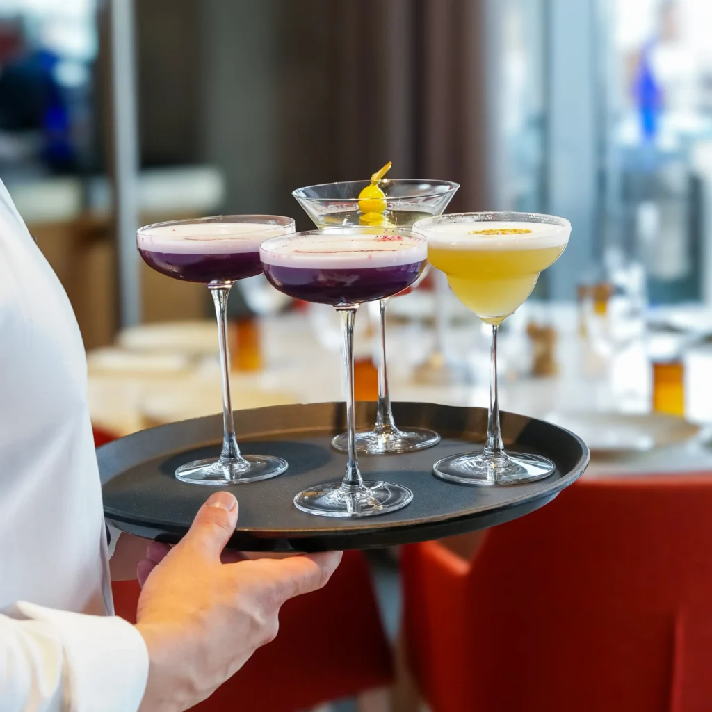 Beyaz üniformalı bir garson, modern bir restoran atmosferinde, zarif ayaklı kadehlerde sunulan ikisi mor renkte köpüklü, biri sarı garnitürlü berrak ve diğeri portakal kabuğu rendesiyle süslenmiş sarı renkli olmak üzere dört çeşit kokteylin bulunduğu siyah bir servis tepsisi taşıyor.
