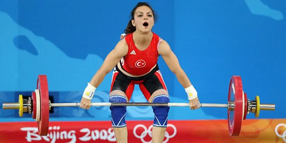 Sibel Özkan, kırmızı halterci mayosu, dizlikleri ve bilek sargılarıyla, ağzı açık, büyük bir güçle Pekin 2008 Olimpiyatları'nda halter kaldırırken, arkada mavi bir fon ve önde Beijing 2008 yazısı ile Olimpiyat halkaları görülüyor.