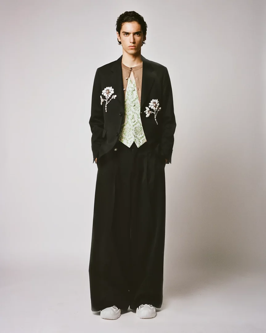 Orta uzunlukta koyu saçlı genç bir model, Simone Rocha'nın Pitti Uomo için tasarladığı romantik ve zanaatkar erkek modası koleksiyonundan, inci işlemeli siyah bir blazer ceket, açık kahverengi hırka, yeşil desenli yelek ve bol paça siyah pantolon giymiş şekilde stüdyoda poz veriyor.