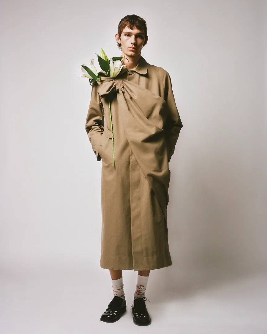 Model, Simone Rocha'nın erkek giyim koleksiyonundan, omuzdan çapraz inen büzgülü ve fiyonk detaylı bej rengi uzun bir palto giyiyor; bu paltonun içinden birkaç beyaz zambak çıkarak Floransa'daki Pitti Uomo'da sergilenecek romantik ve özgün erkek modasına dair güçlü bir ipucu veriyor.
