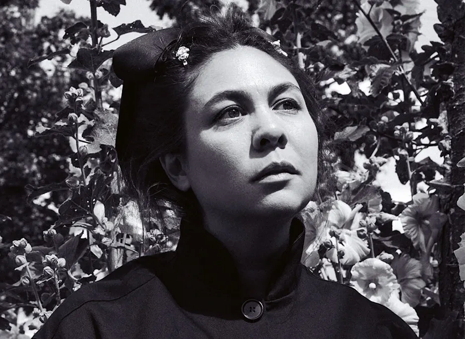 Simone Rocha, Pitti Uomo 110'da erkek modasına yön veren bir konuk tasarımcı olarak, siyah beyaz portresinde hafifçe yukarıya ve sağa bakarken, saçlarında çiçek detayları ve yüksek yakalı koyu renkli bir giysiyle, yeşil bitkilerin ve aydınlık çiçeklerin önünde derin düşüncelere dalmış bir şekilde poz veriyor.