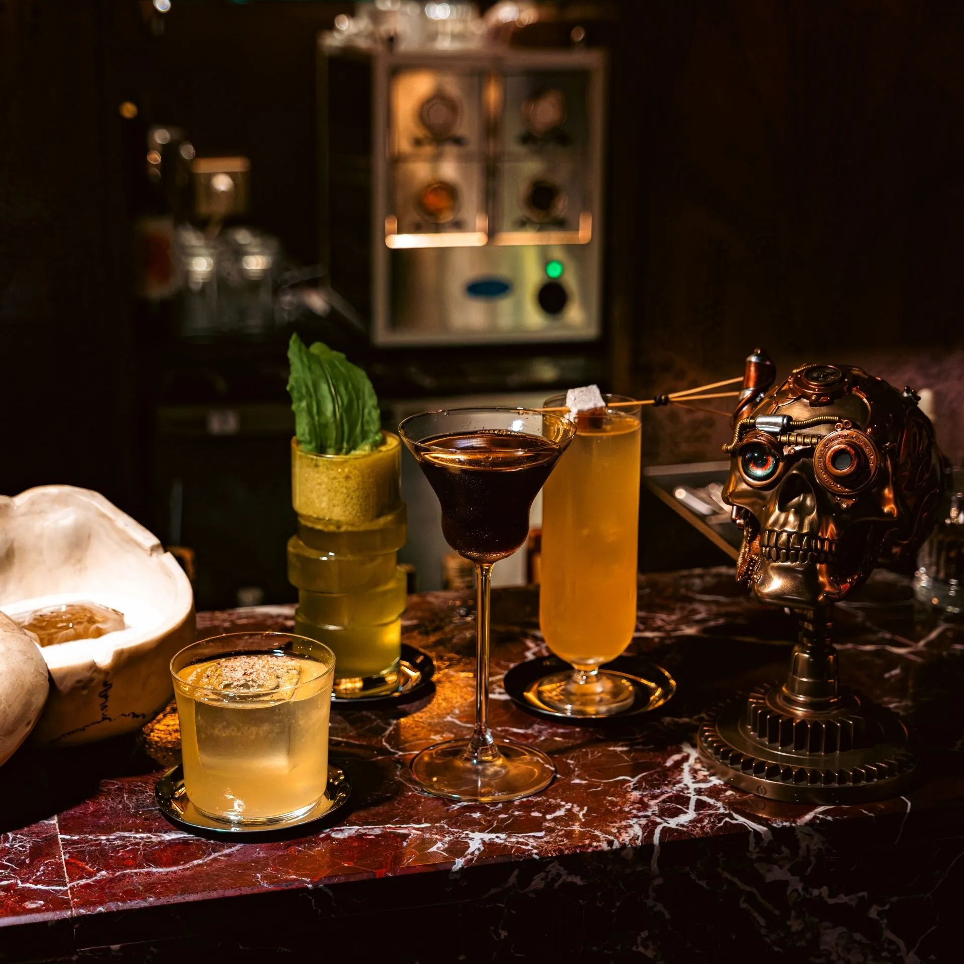 Karaköy'deki loş bir barda, kırmızı mermer tezgah üzerinde koyu renkli bir martini, buz küplü sarı bir içecek, yeşil yapraklı katmanlı bir kokteyl ve steampunk tarzı detaylara sahip metalik bir kurukafa heykeli göz alıcı bir düzenleme sergiliyor.