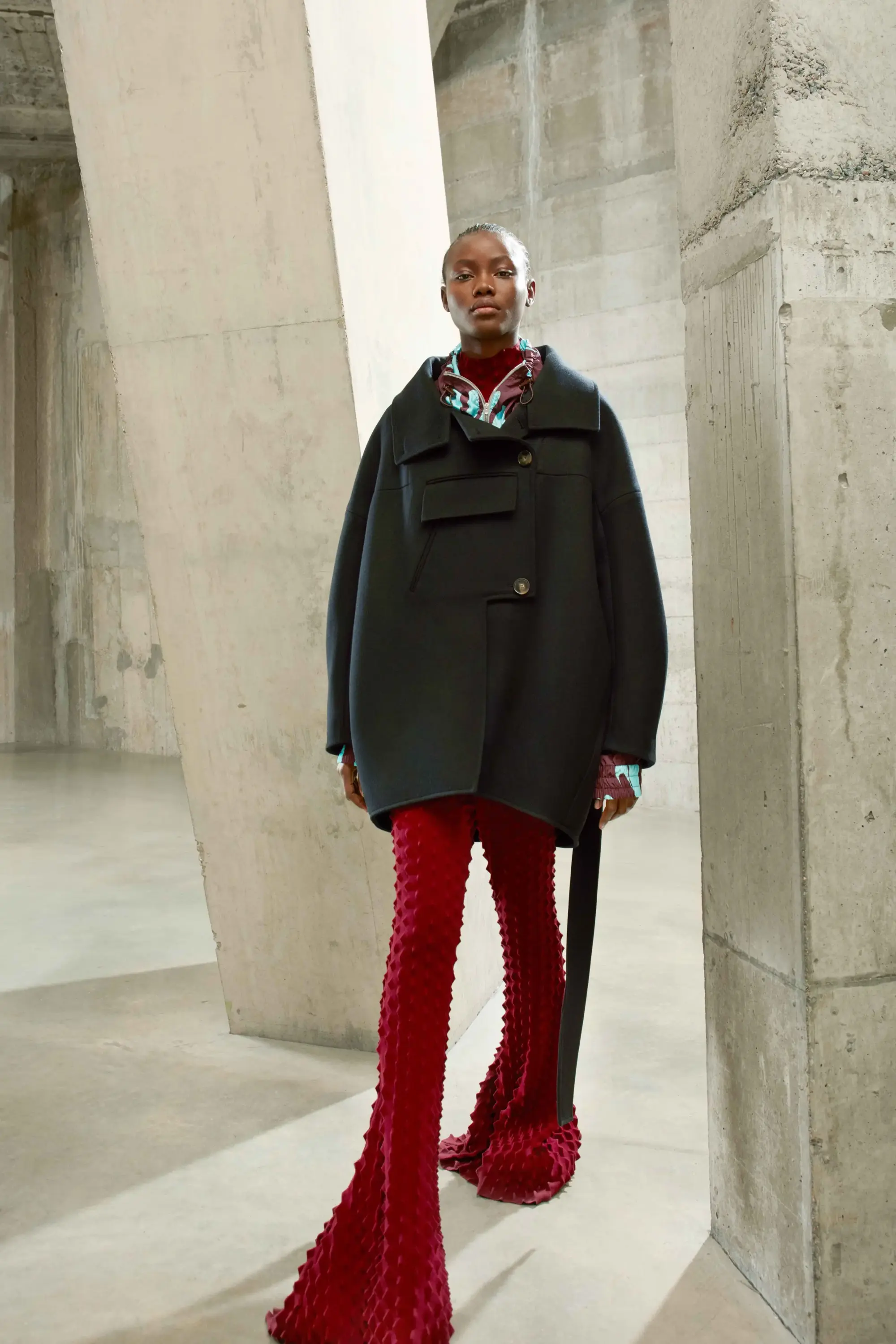 Stella McCartney Sonbahar/Kış 2021 defilesinden siyah oversize palto ve dokulu kırmızı pantolonlarla sürdürülebilir moda estetiği.