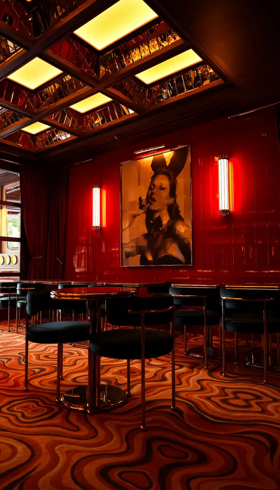 Akaretler'deki modern bir bar veya salonun iç mekanında, kırmızı parlak duvarlar üzerinde tavşan kulaklı bir kadın portresi ve dikey ışıklandırmalar dikkat çekerken, geometrik desenli aynalı tavan ve turuncu tonlarda dalgalı desenli halı, koyu renk sandalyeleri olan yuvarlak masalarla sofistike bir atmosfer yaratıyor.