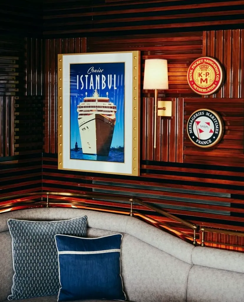 Ahşap panelli şık bir topside bar iç mekanında, Kız Kulesi'nin bulunduğu İstanbul Boğazı'nda bir gemiyi resmeden büyük bir Cruise İstanbul posteri, Royal Packet Navigation ve Messageries Maritimes gibi tarihi denizcilik firmalarının amblemleri ile birlikte pirinç aplikle aydınlatılmış ahşap duvarda yer alıyor ve altında kıvrımlı, döşemeli bir kanepe ve minderler görülüyor.