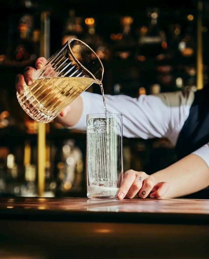 Barmen, loş ışıklı şık bir bar tezgahında, elindeki çizgili sürahiden köpüklü, açık sarı renkli bir kokteyli uzun, şeffaf bir bardağa dikkatlice dolduruyor, arka planda bulanık şişeler ve ışıklar görünüyor.