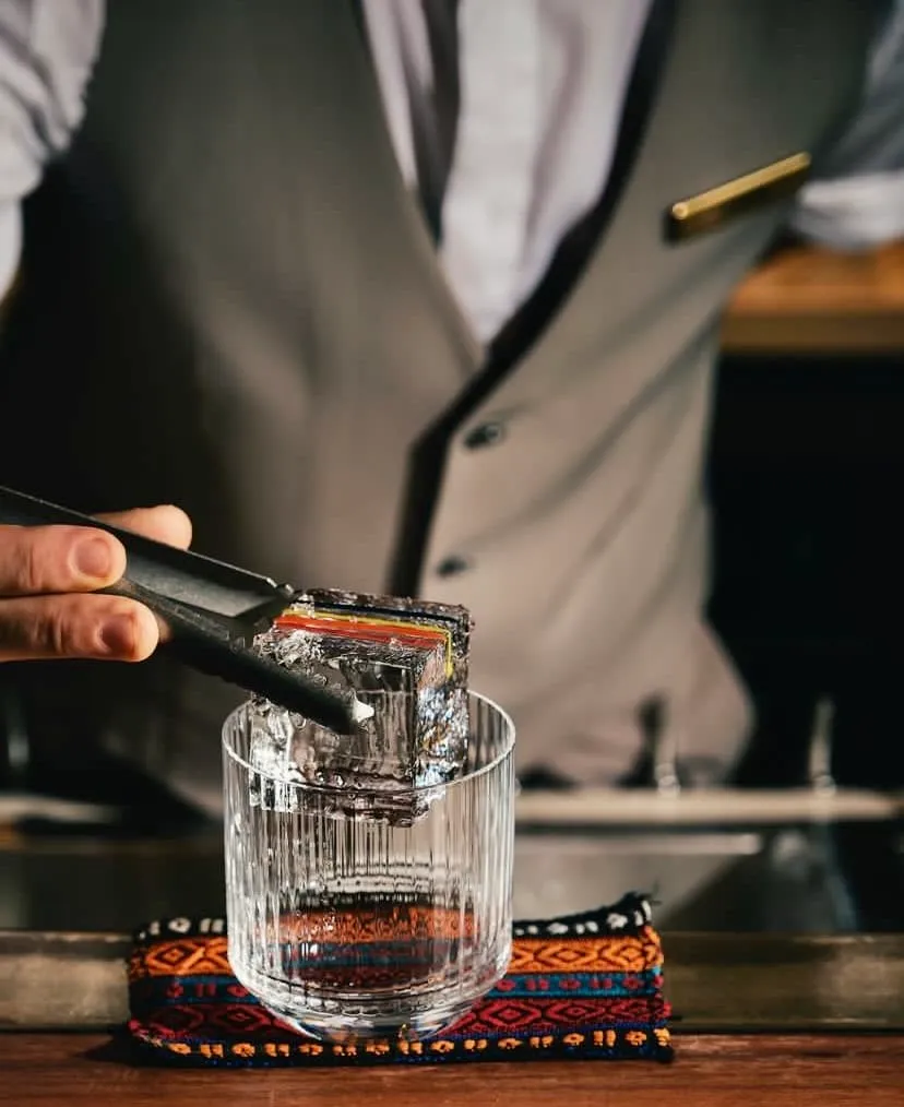 Gri yelekli bir barmen, bar tezgahındaki renkli desenli bir nihale üzerindeki çizgili şeffaf bardağa, üst kenarında belirgin ince renkli katmanları olan büyük, kristal netliğindeki bir buz küpünü buz maşasıyla özenle bırakıyor.