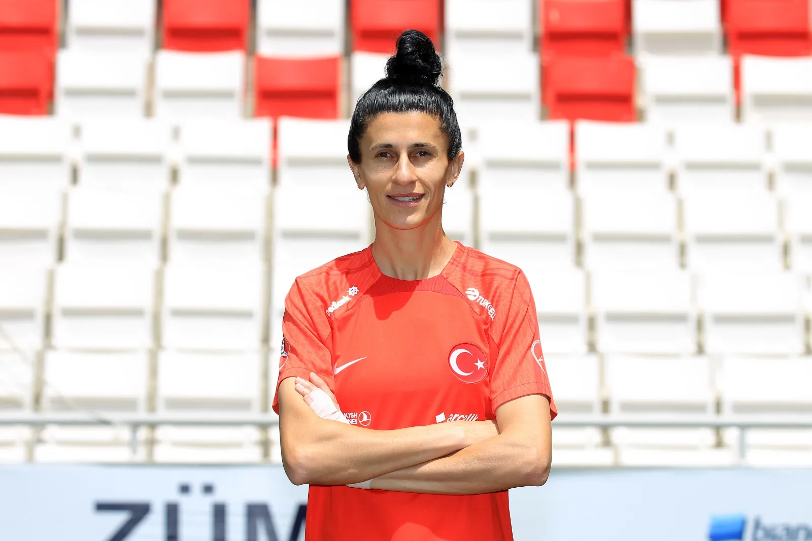 Sol bileğinde beyaz bandaj bulunan kadın milli futbolcu Yağmur Uraz, üzerinde Türk bayrağı ve sponsor logoları olan kırmızı formasıyla gülümsüyor, saçları topuz yapılmış ve kolları bağlı bir şekilde, arka planda kırmızı ve beyaz koltuklu bir stadyum tribünü önünde poz veriyor.