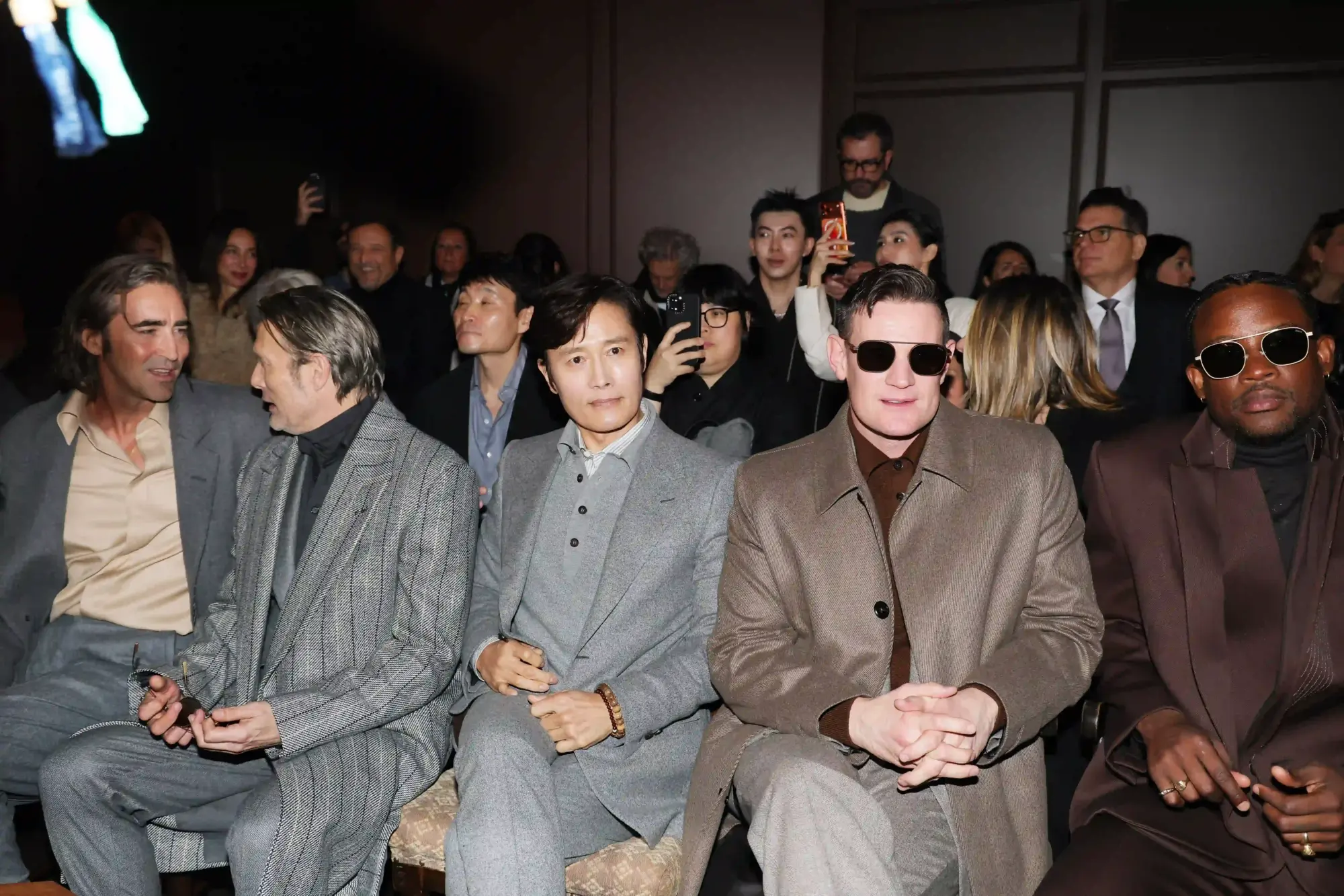 Zegna 2026 defilesi front row’da gri ve kahverengi tonlarda klasik takım elbise ve paltolarla oturan davetliler, modern erkek giyimi şıklığını yansıtan moda haftası atmosferi.