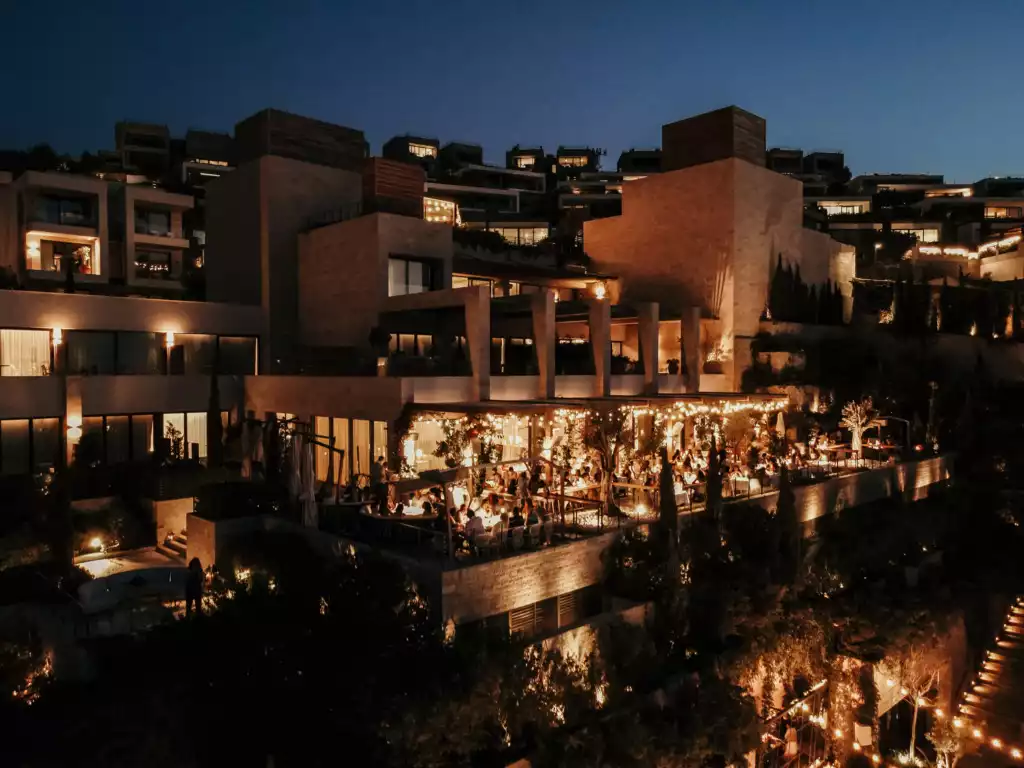 Bodrum Edition otelinin gece çekilmiş bu geniş açılı fotoğrafında, taş cepheli katmanlı mimarisi, ışıl ışıl parlayan pencereleri ve ışık seli altındaki açık hava restoranında keyifli anlar geçiren çok sayıda misafiriyle adeta bir şölen sunuluyor.